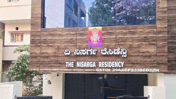 El exterior de The Nisarga Residency, con una fachada moderna, una amplia puerta de entrada y vista a la calle.
