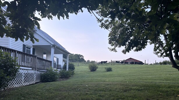 Ein Haus mit Veranda, einem eingezäunten Garten und einem Bauernhof im Hintergrund.