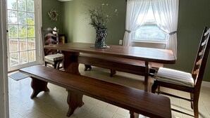 Ein Essbereich mit Holztisch und Bänken, einer Blumenvase und einem mit Vorhängen drapierten Fenster.