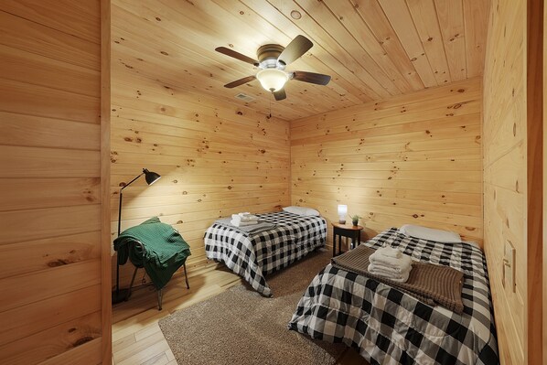 A wooden cabin with two beds, a ceiling fan, a lamp, and a small table with a lamp.