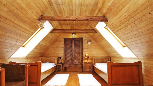 Un dormitorio cálido en el ático con paredes de madera y techos inclinados, con dos camas individuales, una silla de mimbre y una mesita pequeña.