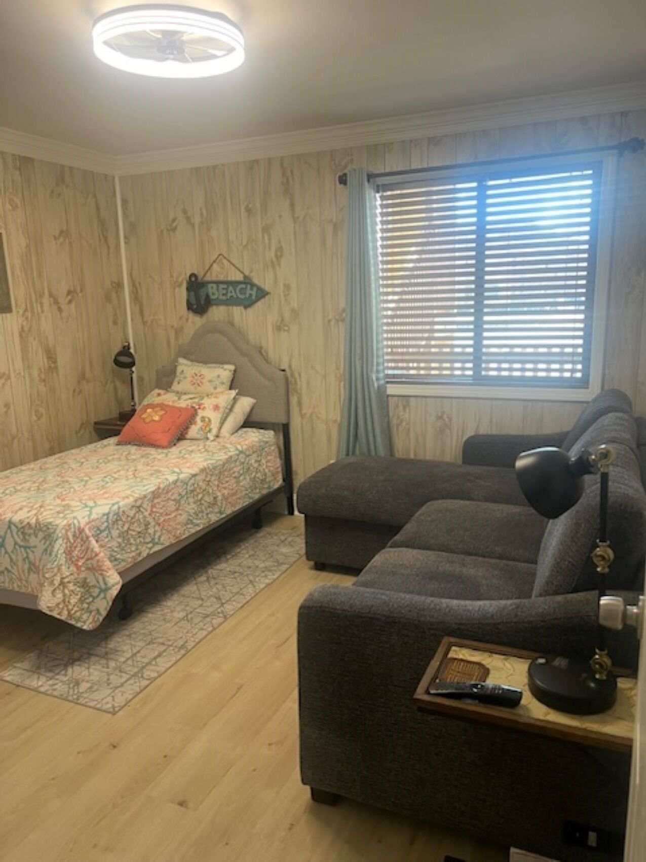 Ett sovrum med en säng, en soffa, ett skrivbord, en lampa, ett fönster med persienner och en skylt på väggen.