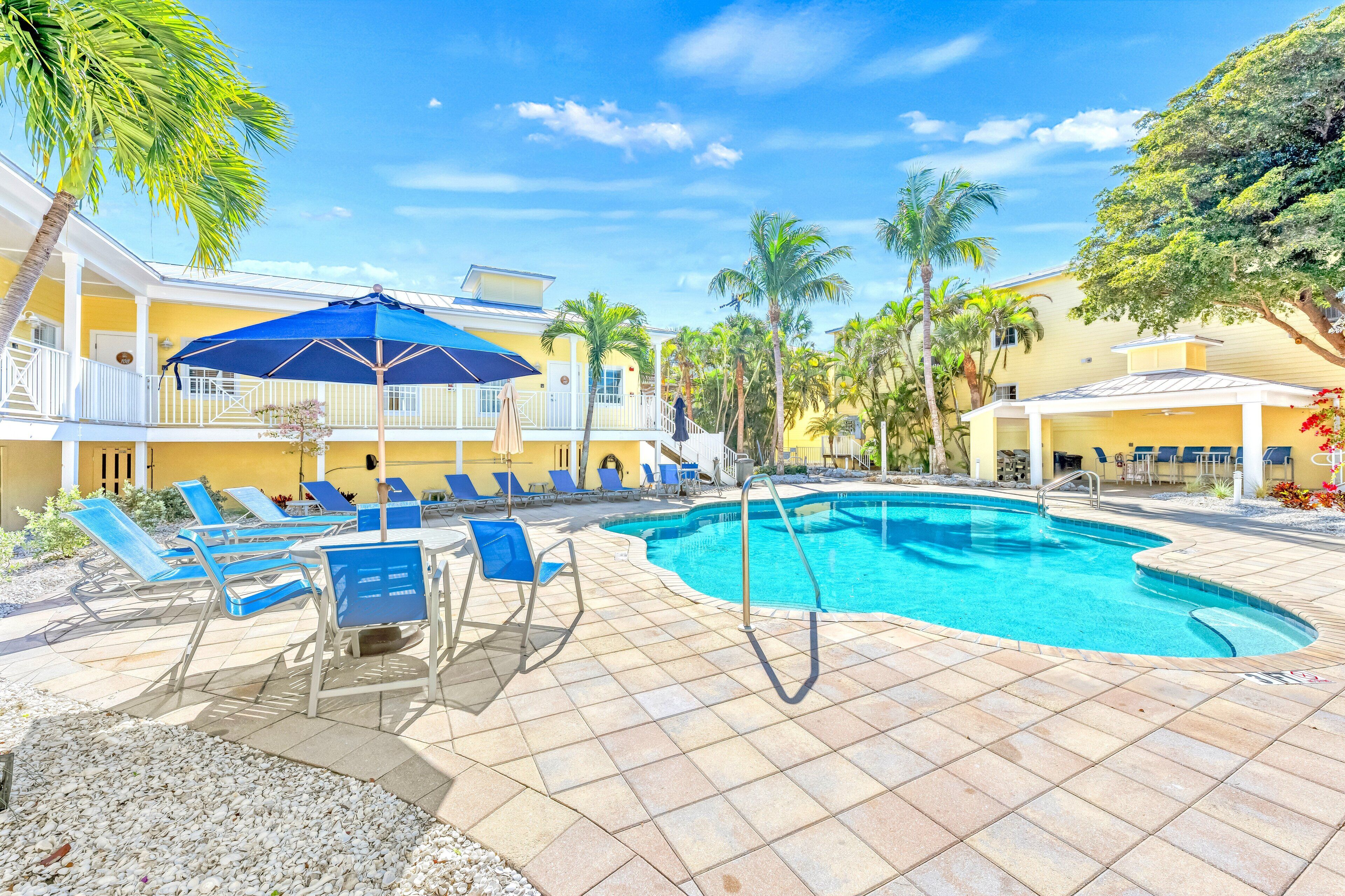 A pool area with lounge chairs and umbrellas, surrounded by palm trees and a building with balconies.