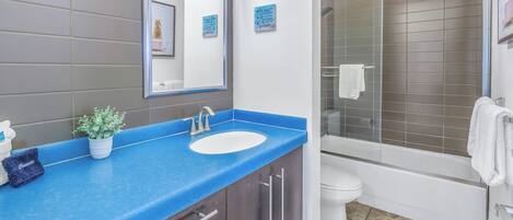 Une salle de bain avec un plan de toilette bleu, un lavabo blanc, un miroir, une plante en pot, une douche et des toilettes.