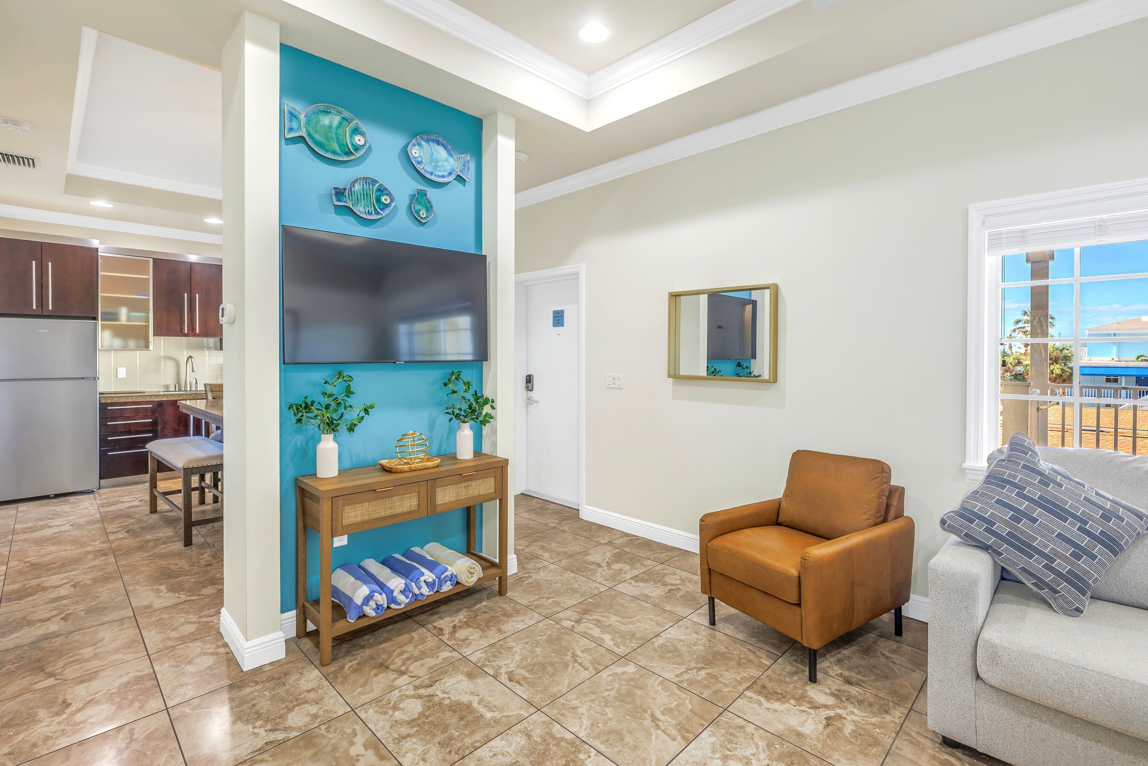 A spacious living area with a sofa, a coffee table, and a TV. There is a kitchen in the background with cabinets and a refrigerator.