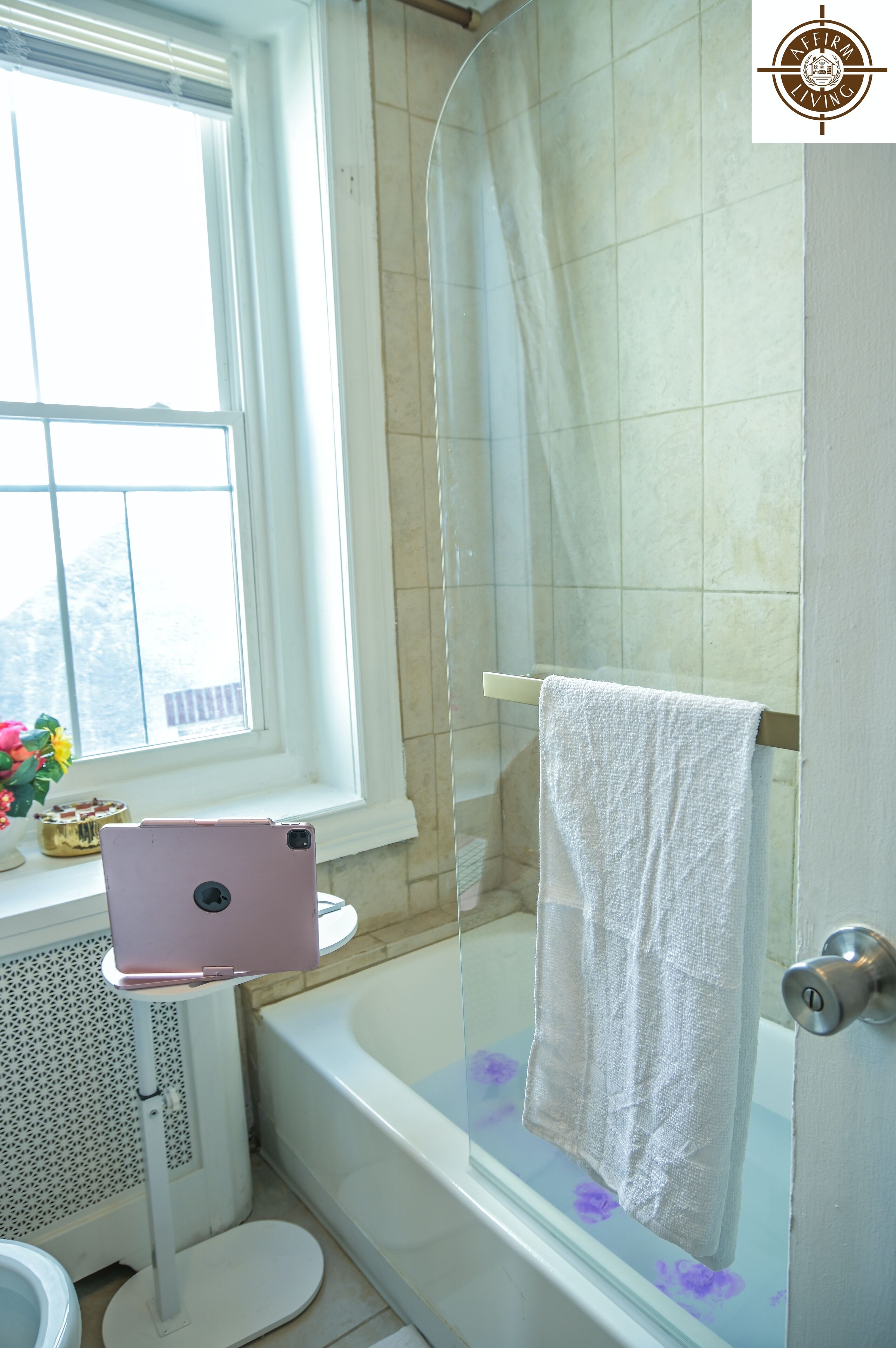 Een badkamer met een glazen douchecabine, een wit bad en een witte handdoek aan de douchedeur, plus een roze tablet op een standaard.