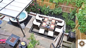 Een terras met een tafel, stoelen, een hottub en een houten omheining begroeid met klimplanten.