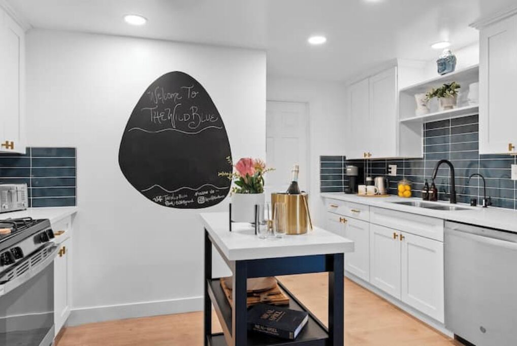 A modern kitchen with white cabinetry, a black chalkboard wall with writing, and a small table with decorative items.