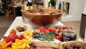 Una tabla de fiambres con varios quesos, frutas y embutidos, sobre una mesa de madera en una cocina moderna con una pared de ladrillo blanco y una chimenea al fondo.