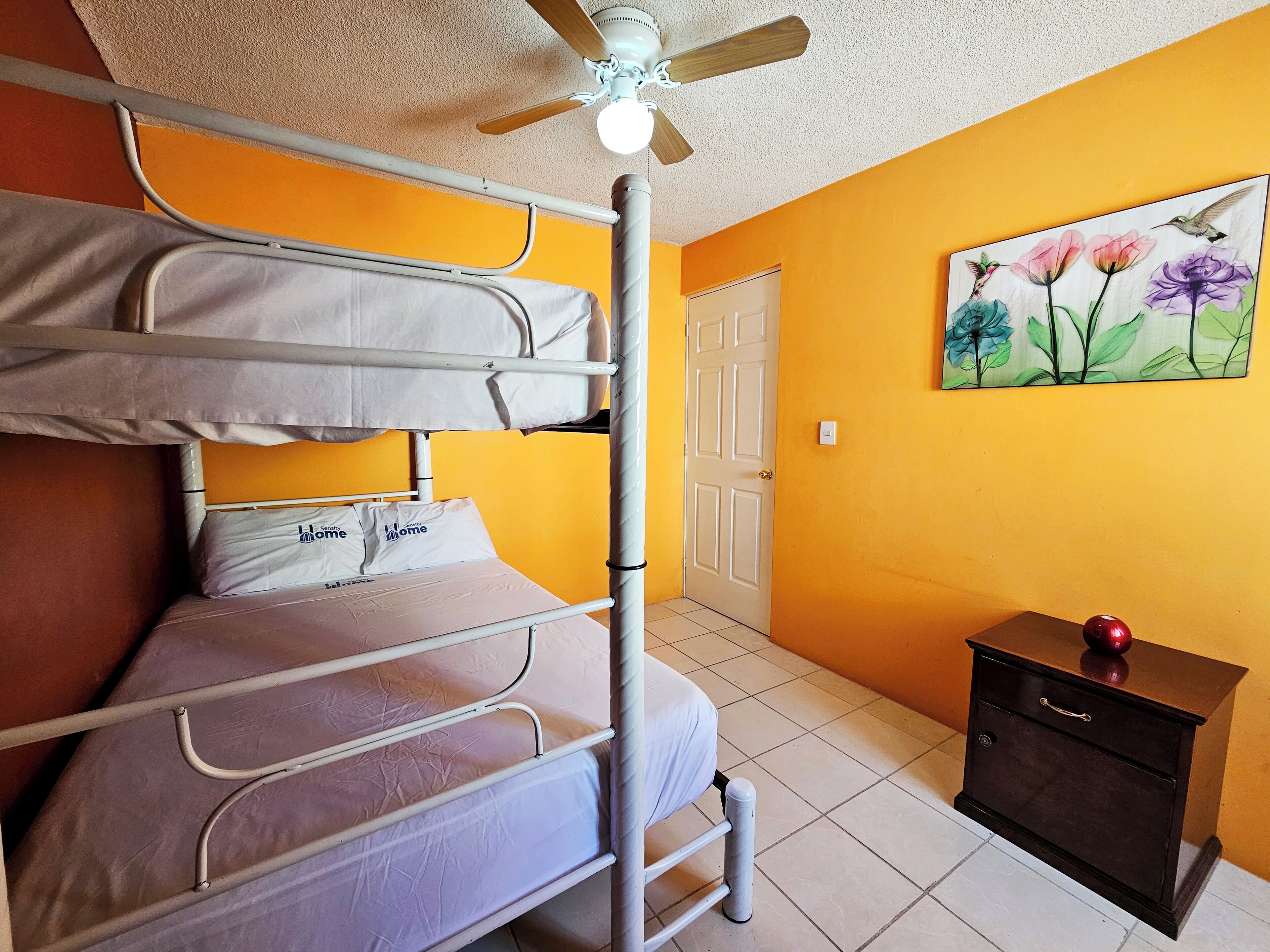 A room with a bunk bed, a ceiling fan, a door, a painting, and a small wooden cabinet.