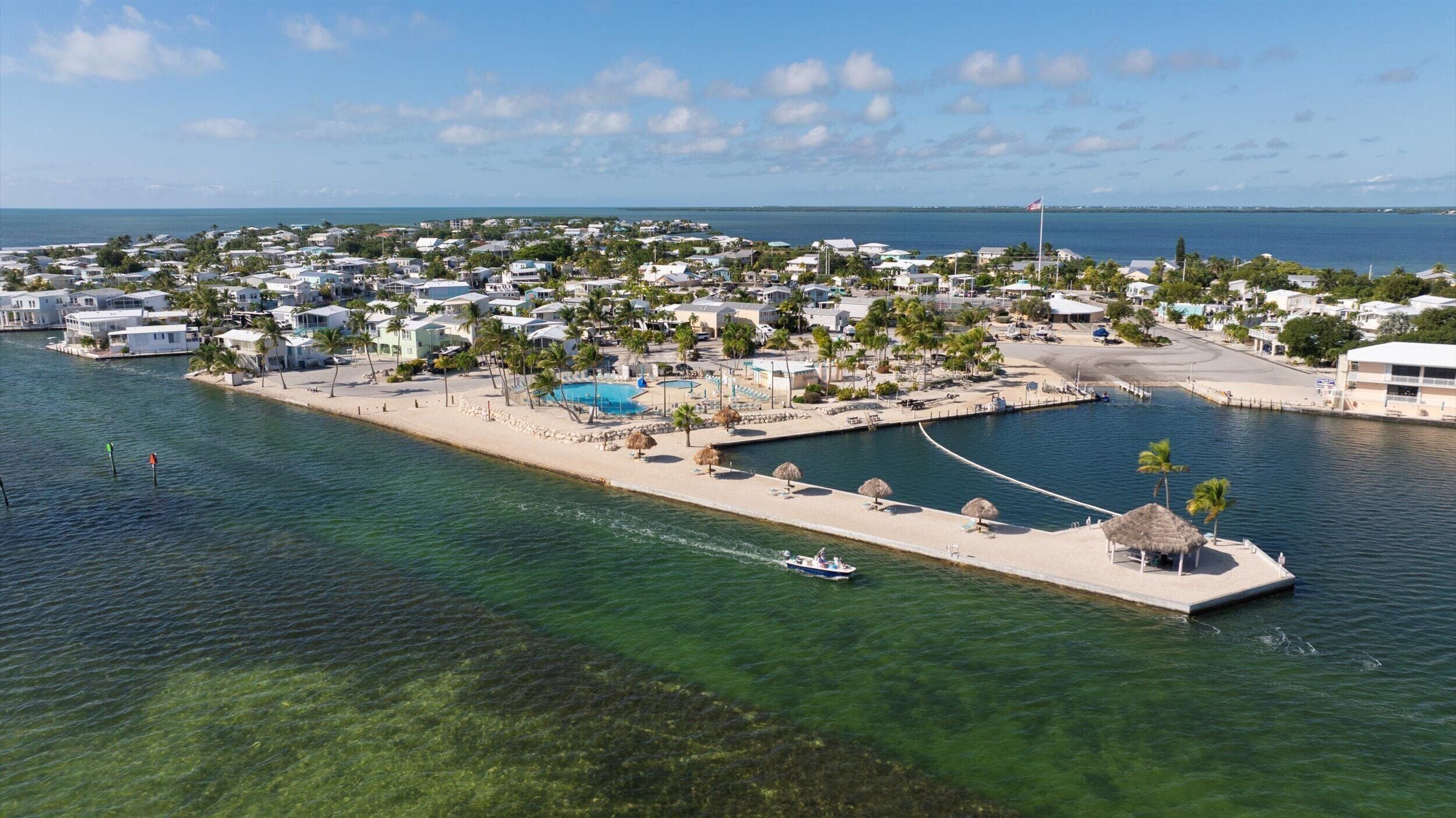 Ett flygbildsbild av en kustort med en småbåtshamn, klart vatten och ett strandområde med parasoller och en swimmingpool.