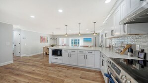 A modern kitchen with white cabinetry, a central island, and a built-in oven. There are pendant lights above the island and a backsplash with a mosaic tile design. - Heated Pool Retreat • 3BR • Minutes to Siesta Key & Downtown (Sarasota)