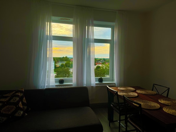Una sala de estar con un sofá, una mesa de comedor con individuales y vistas a un atardecer a través de una ventana.
