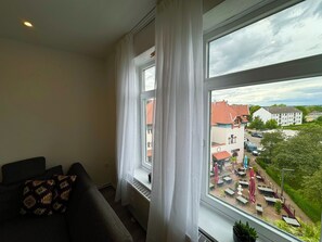 Una habitación con un ventanal grande, un sofá oscuro con cojines estampados, y vistas a un pueblo con edificios y un área verde.