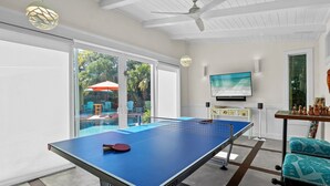 A ping pong table in a room with a TV, a ceiling fan, and a view of a pool and palm trees.