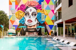 Un área de piscina con un mural, sillones de descanso y un edificio de fachada geométrica.
