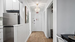 Ein schmaler Flur mit Holzboden, weißen Wänden und einer Chandelierleuchtre. Ein Ausgangeschild ist auf der Tür sichtbar.