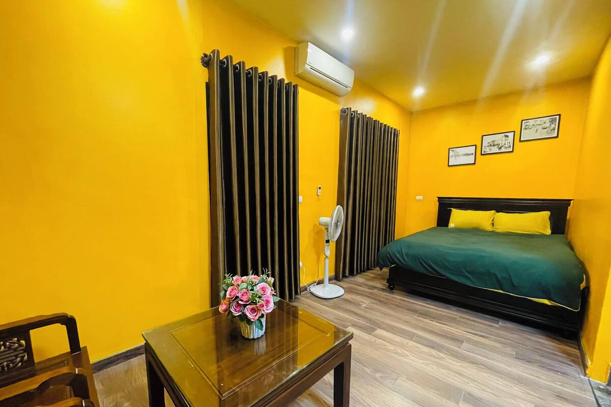 A hotel room with a bed, a table with a vase of flowers, a standing fan, and a window with curtains.
