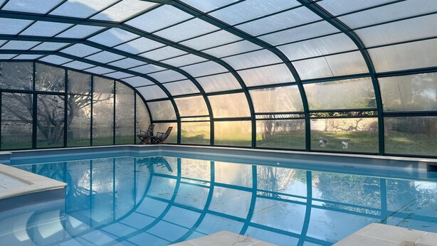 Une piscine intérieure dotée d’un toit en verre, d’une eau d’un bleu cristallin et offrant une vue sur un jardin extérieur.