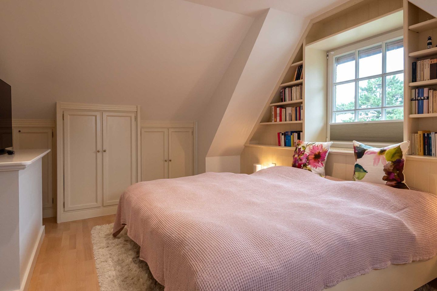 A bedroom with a sloped ceiling, a bed with a pink blanket, a white rug, a bookshelf, and a window with daylight.