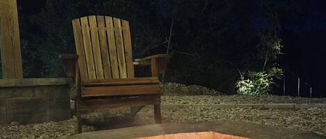 A wooden chair near a fire pit with a stone fire ring, set against a dark night sky and surrounded by trees.