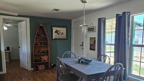 Een eetruimte met een houten tafel en stoelen, een plafondventilator en uitzicht op het landschap door een raam met gordijnen.