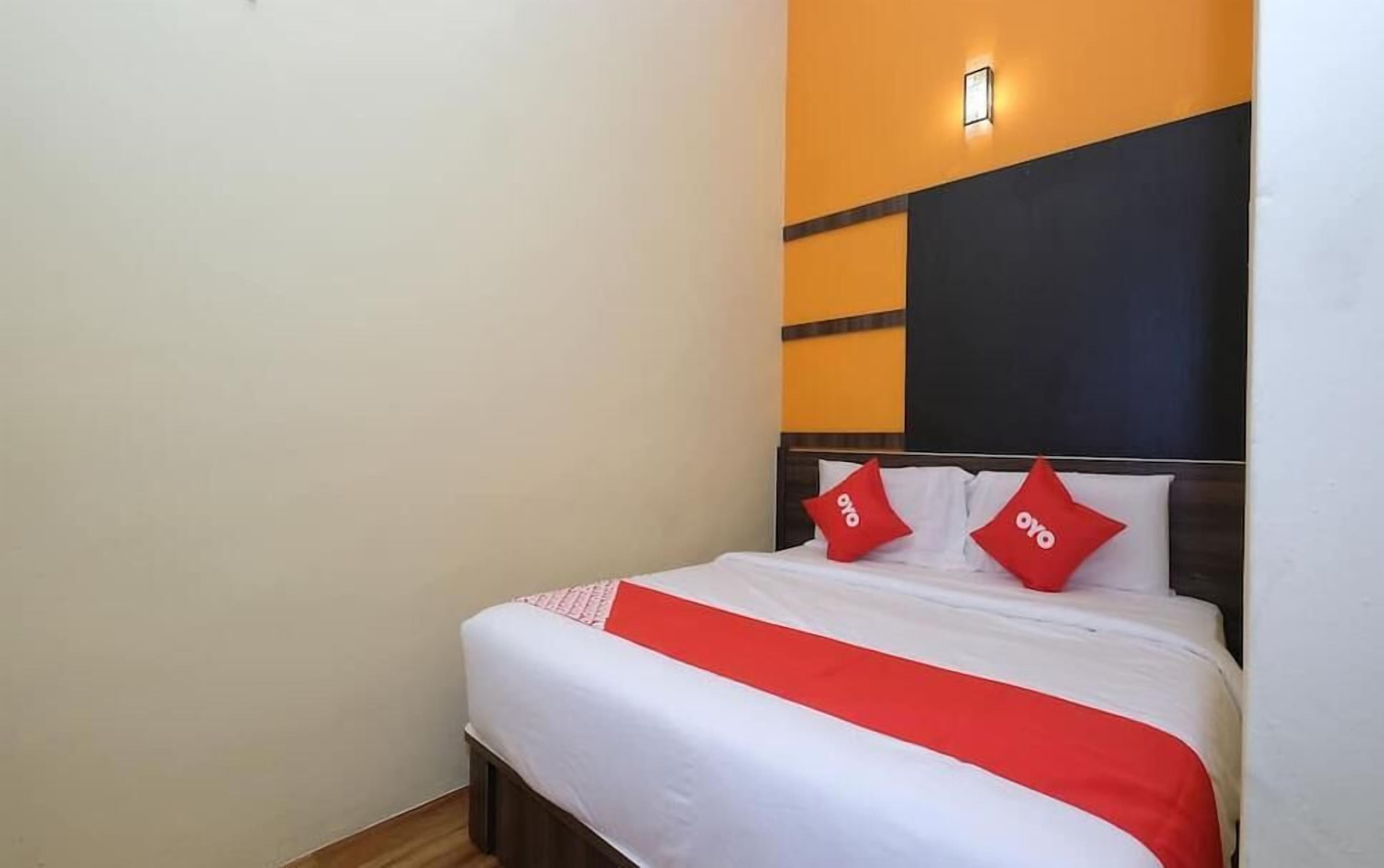 Habitación de hotel con cama, dos almohadas, colcha roja y blanca y una pared con lámpara.