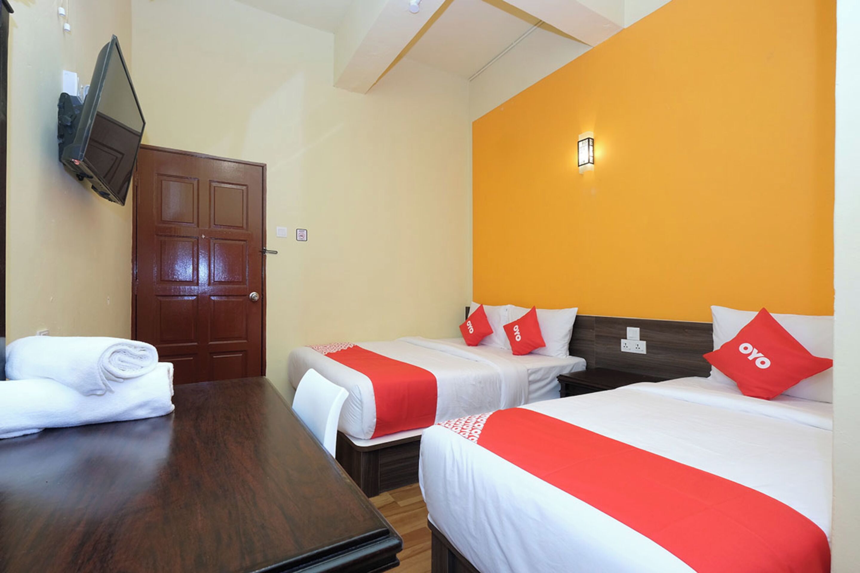Een hotelkamer met twee bedden, een houten tafel, een aan de muur bevestigde televisie en een deur.