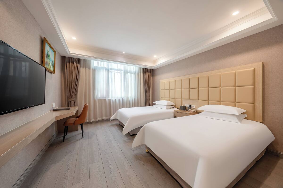 A hotel room with two beds, a desk, a chair, a TV, and a large window with curtains.