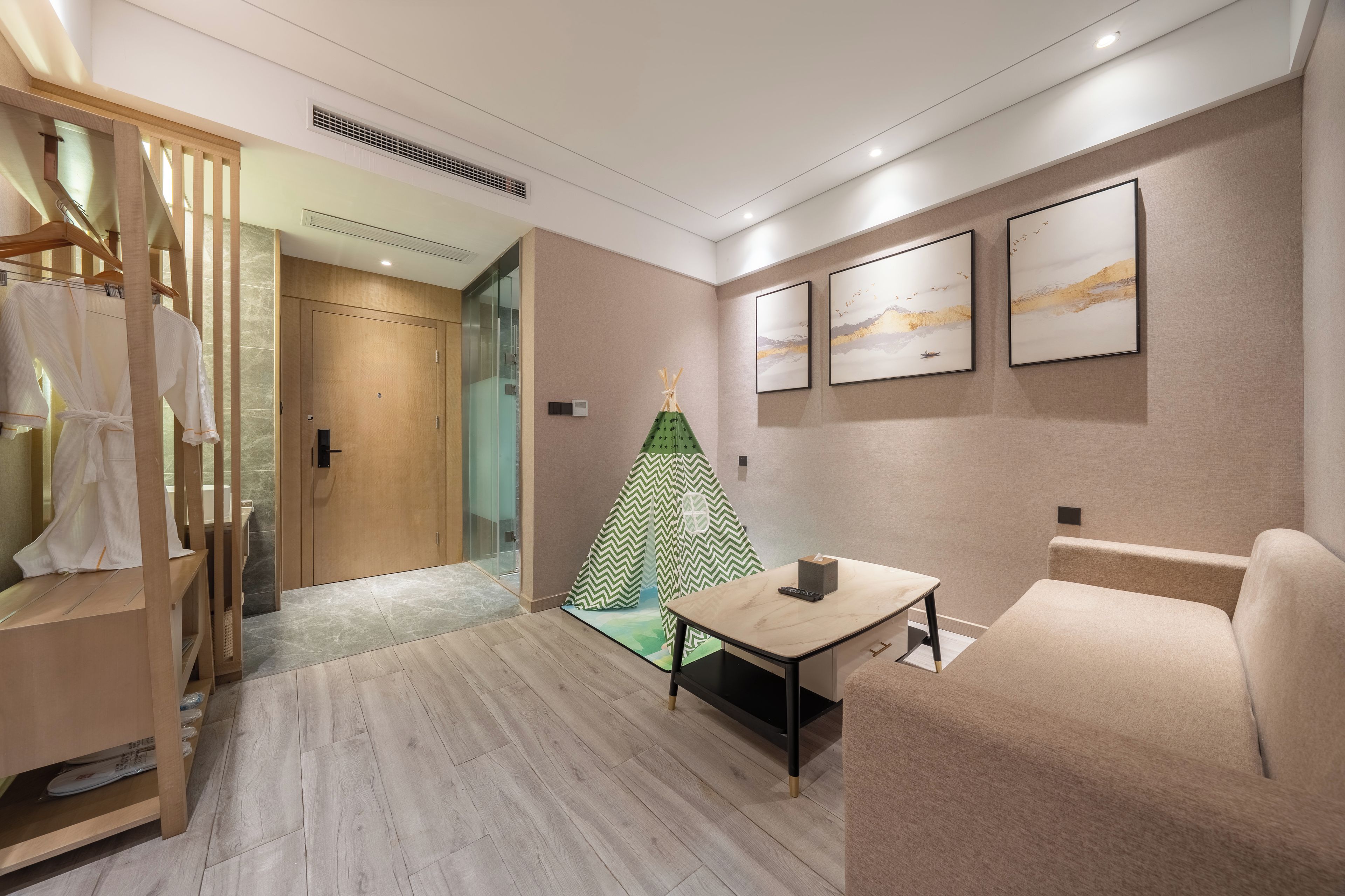 Une chambre d’hôtel moderne avec une armoire en bois, une tente indienne verte et blanche, un canapé beige, une petite table et des œuvres d’art encadrées accrochées au mur.