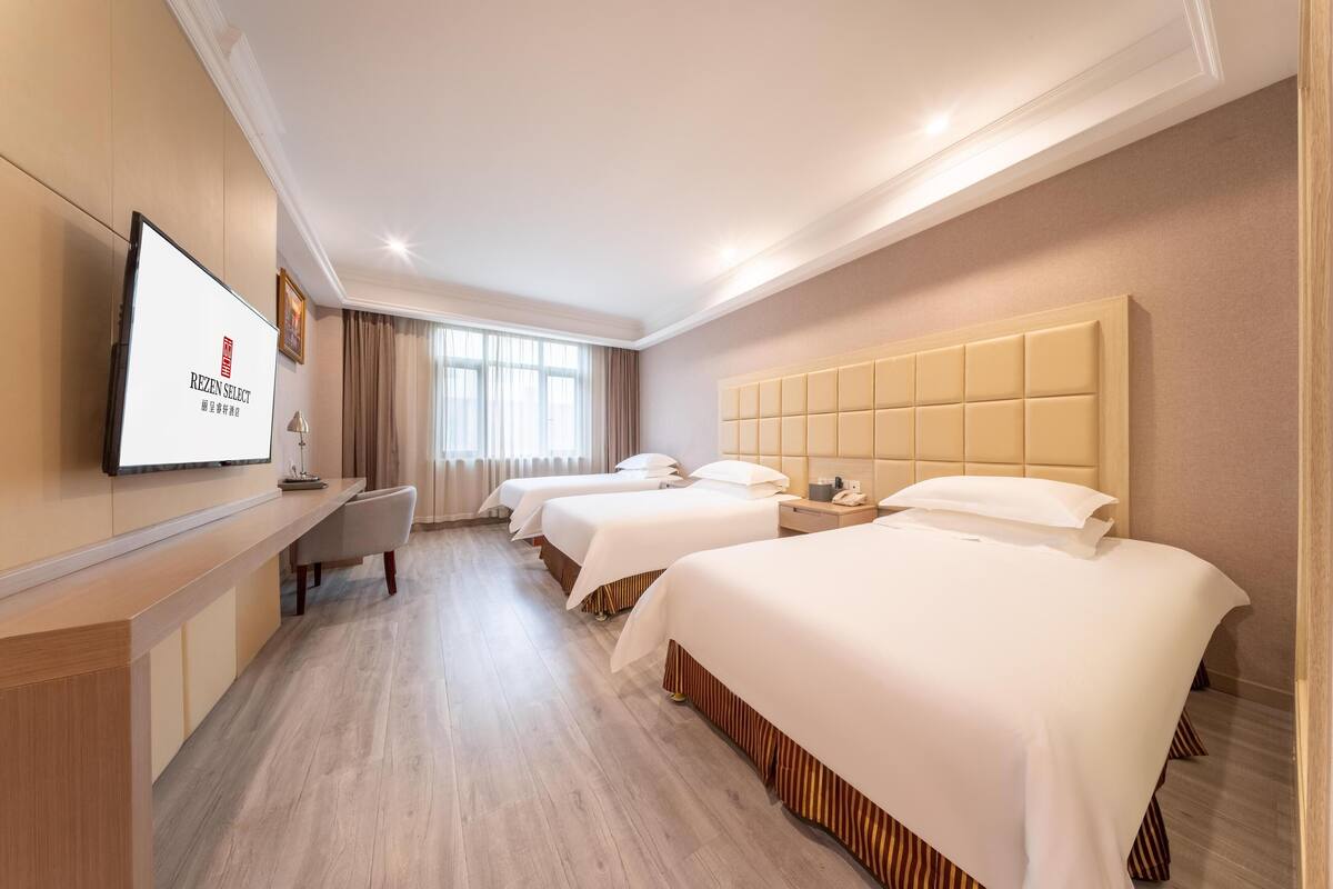 A hotel room with three beds, a desk, a television, and a large window with curtains.