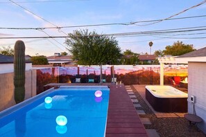 A swimming pool with hot tub, surrounded by a deck with seating and a view of a sunset.