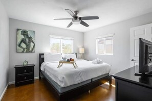 A bedroom with a bed, a nightstand, a desk, a ceiling fan, and a painting on the wall.