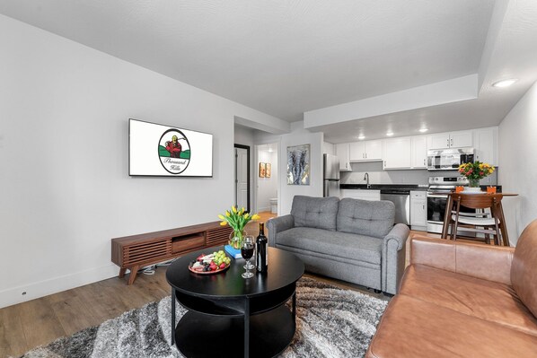Een moderne woonkamer met een grijze bank, een zwarte salontafel, en een eethoek met een houten tafel en stoelen.