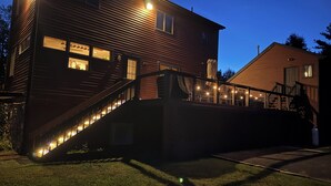 Una casa de dos pisos con una escalera que lleva al nivel superior, un porche adornado con luces colgantes y una puerta de garaje visible.