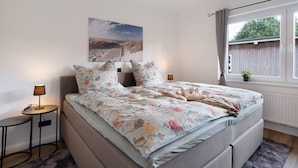 Un dormitorio con cama, mesita de noche, lámpara, ventana con cortinas e imagen colgada en la pared.