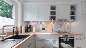 Una cocina moderna con armarios blancos, un salpicadero con diseño, horno empotrado y una ventana con persianas.