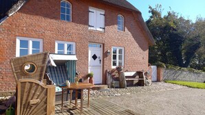 Une maison en briques avec un toit de chaume, une terrasse en bois et un petit espace extérieur aménagé pour s’asseoir.