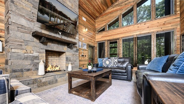 A cozy interior with a stone fireplace, leather furniture, and large windows. - Luxury Cabin*Hot Tub*Fire Pit*Game Room*Arcade (Broken Bow)