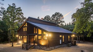A two-story log cabin with a metal roof, a stone foundation, and a gravel driveway. - Luxury Cabin*Hot Tub*Fire Pit*Game Room*Arcade (Broken Bow)