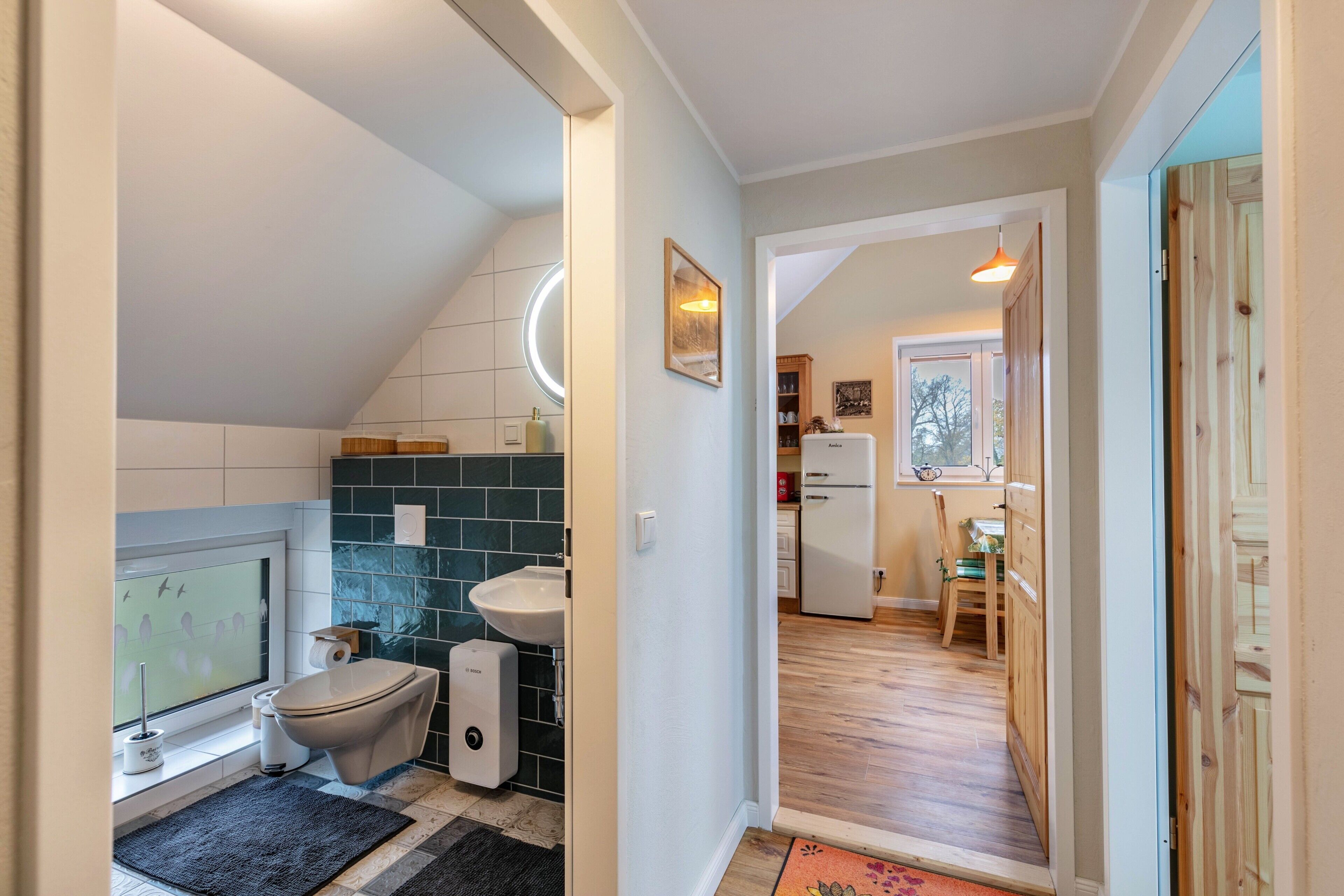 Ein modernes Badezimmer mit Toilette, Bidet und Waschbecken, einem kleinen Fenster mit Aussicht und einem Holzboden, der zum Küchenbereich führt.