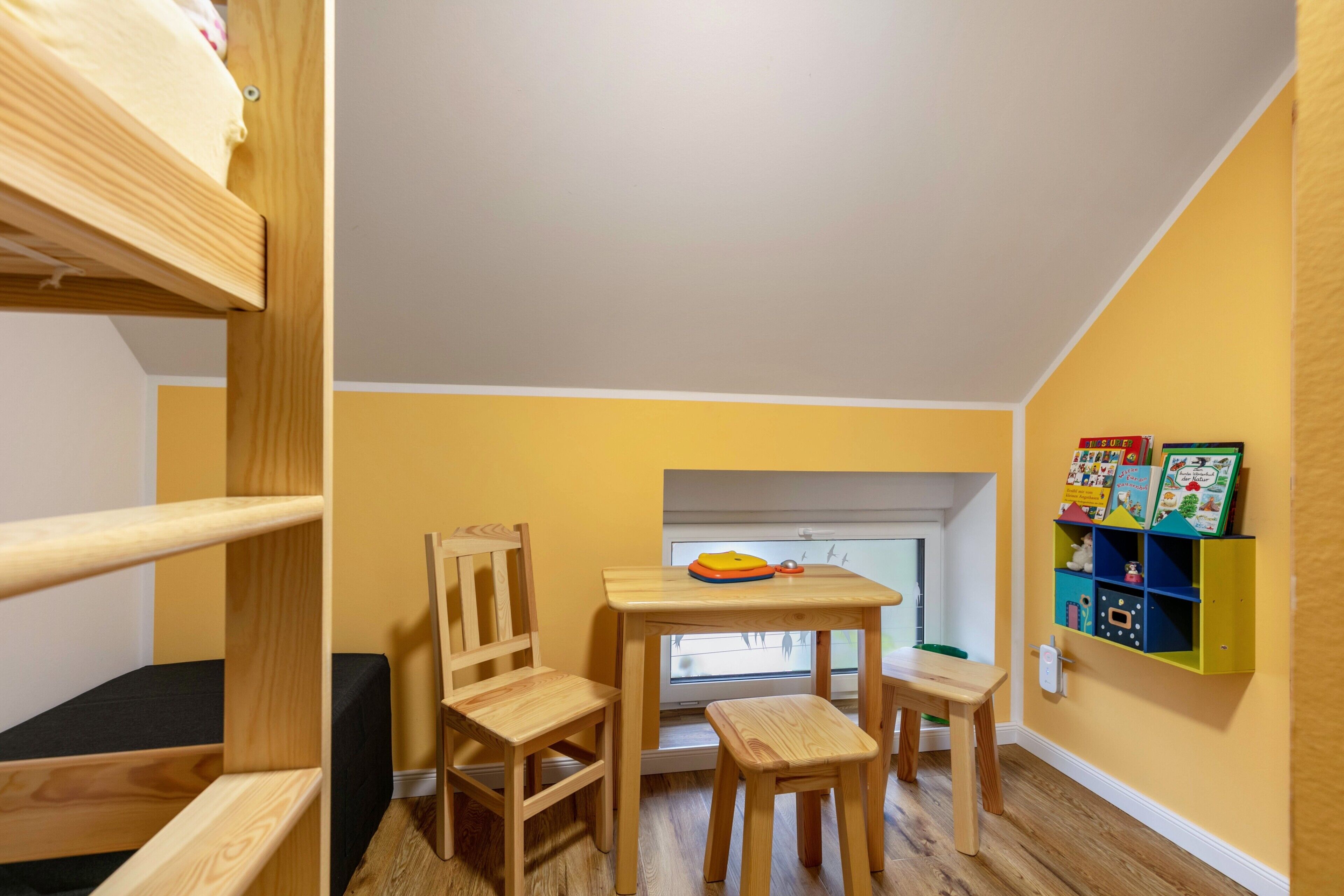 Ein Zimmer mit Etagenbett, einem Holztisch mit Stühlen, einem Fenster und einem Bücherregal mit Kinderbüchern.
