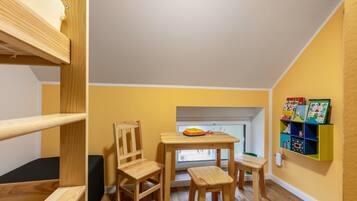 Ein Zimmer mit Etagenbett, einem Holztisch mit Stühlen, einem Fenster und einem Bücherregal mit Kinderbüchern.
