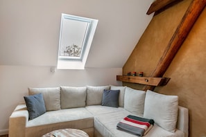 A corner room with a sloped ceiling, a built-in wooden shelf, a sectional sofa with cushions, and a small window.