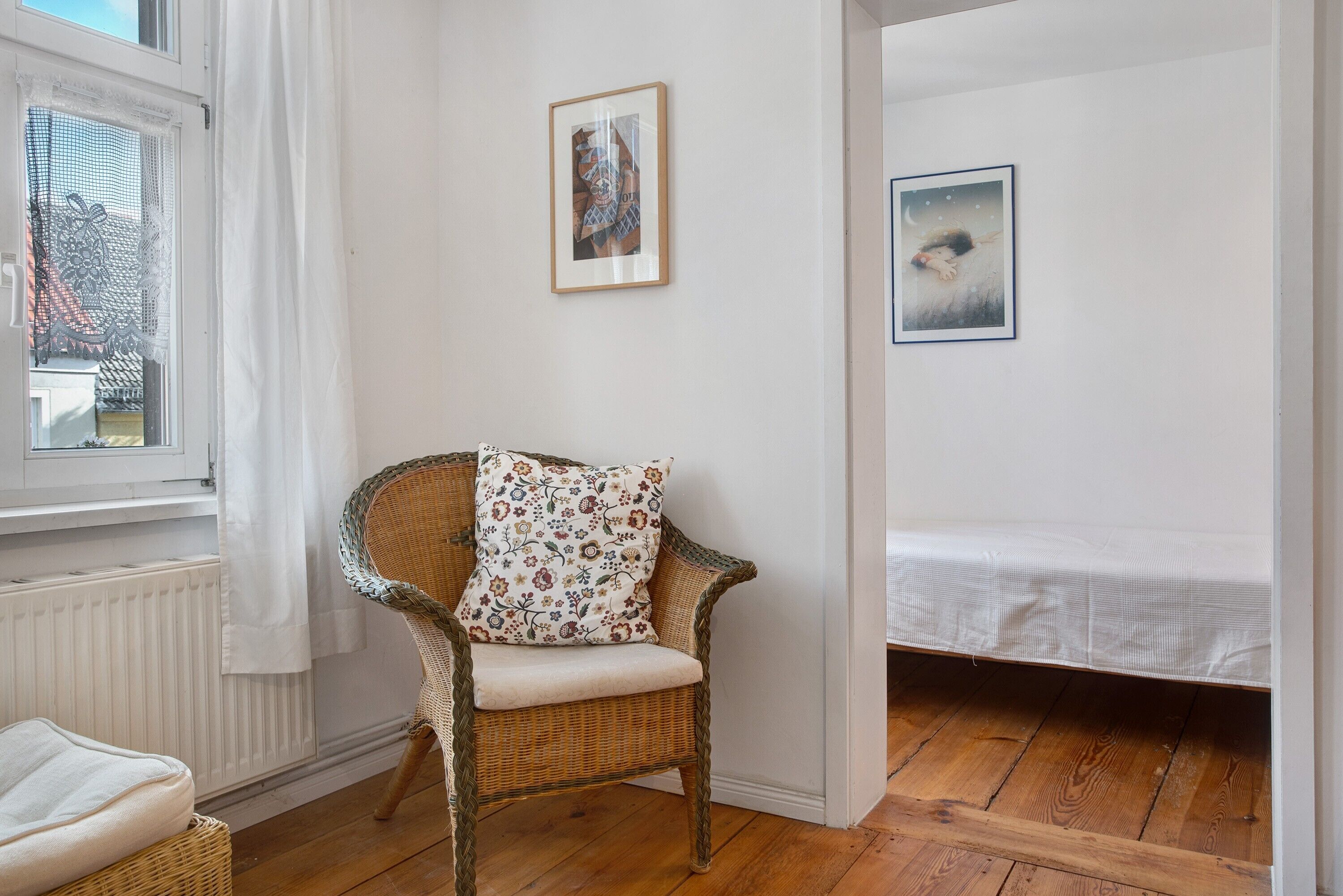Ein Zimmer mit einem Korbstuhl, einem Bett und zwei gerahmten Bildern an der Wand.