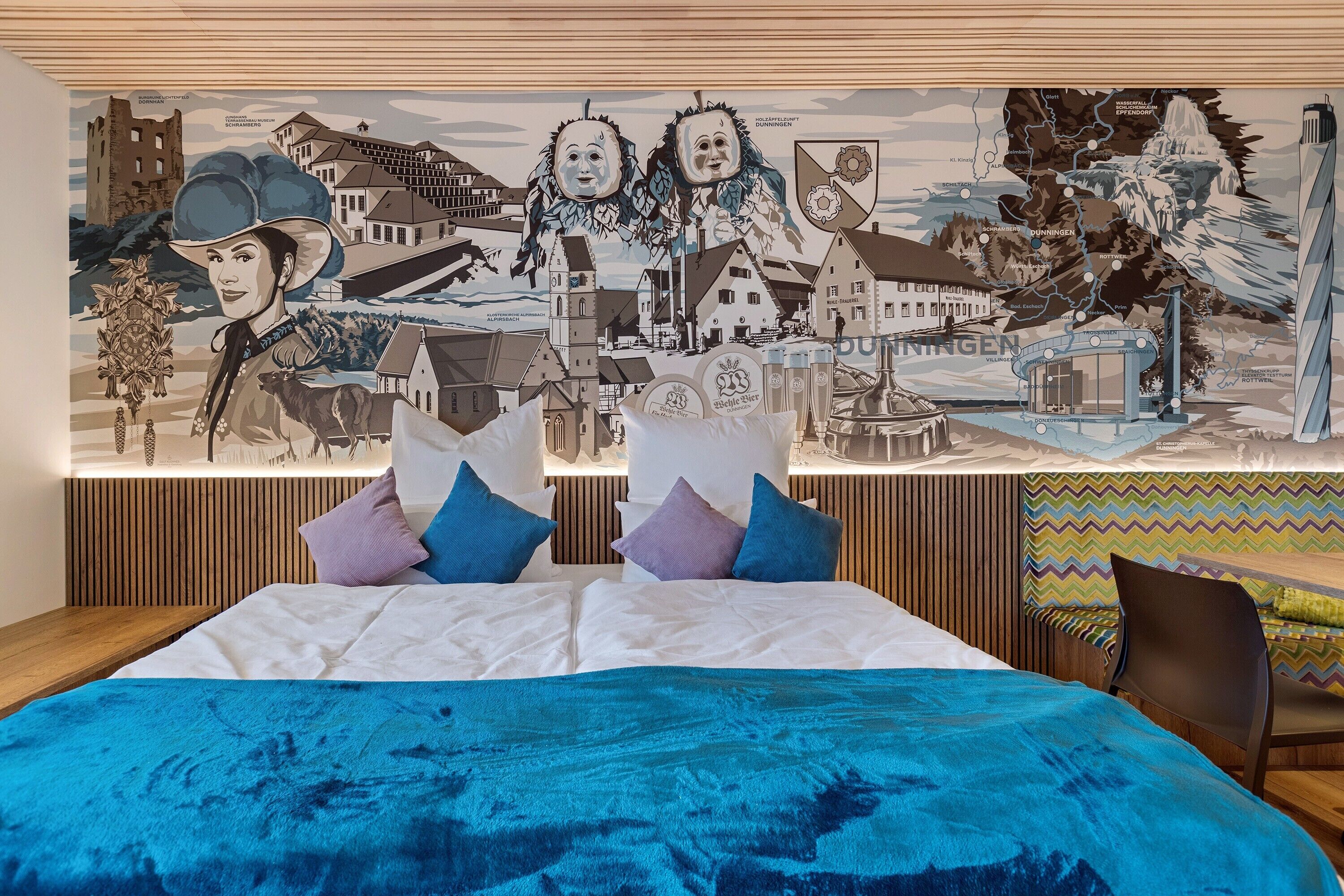 Une chambre d’hôtel avec une grande fresque murale, un lit recouvert d’une couverture bleue et une tête de lit en bois.