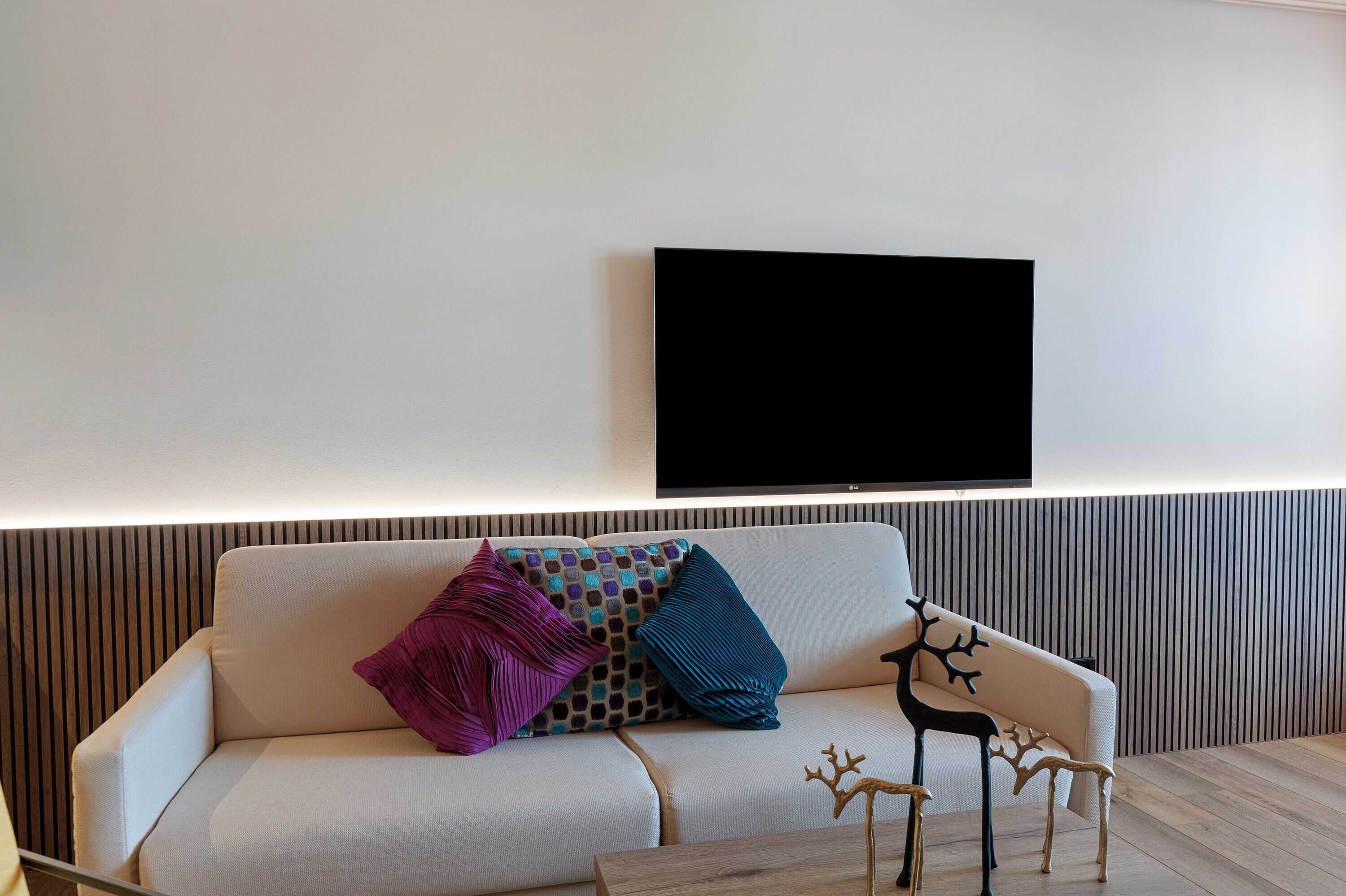 Un salon moderne avec un canapé, des coussins décoratifs, un téléviseur fixé au mur et une table basse en bois.
