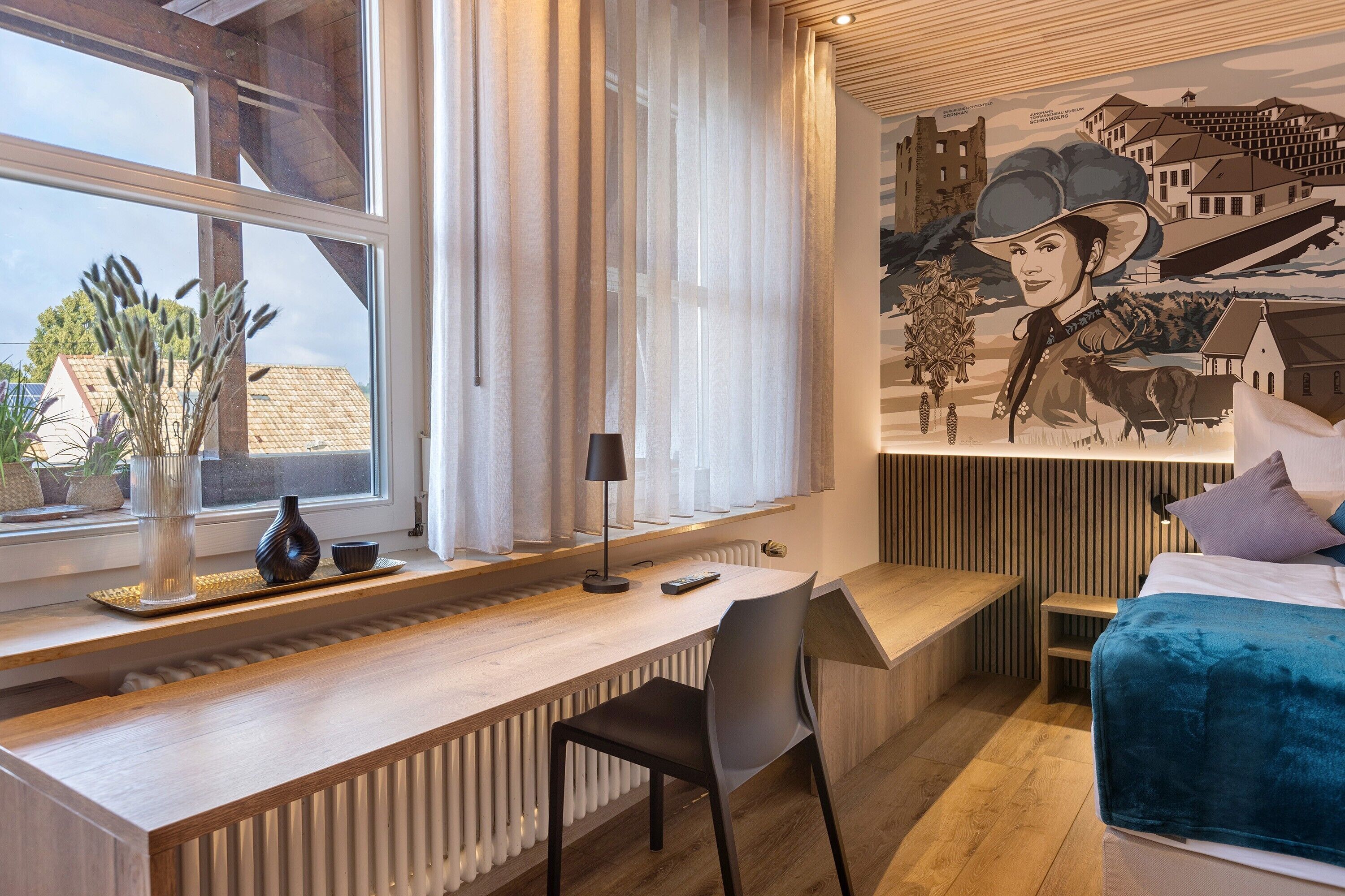 Un dormitorio moderno con un escritorio de madera, una cama con manta azul, un mural y una ventana con cortinas traslúcidas.