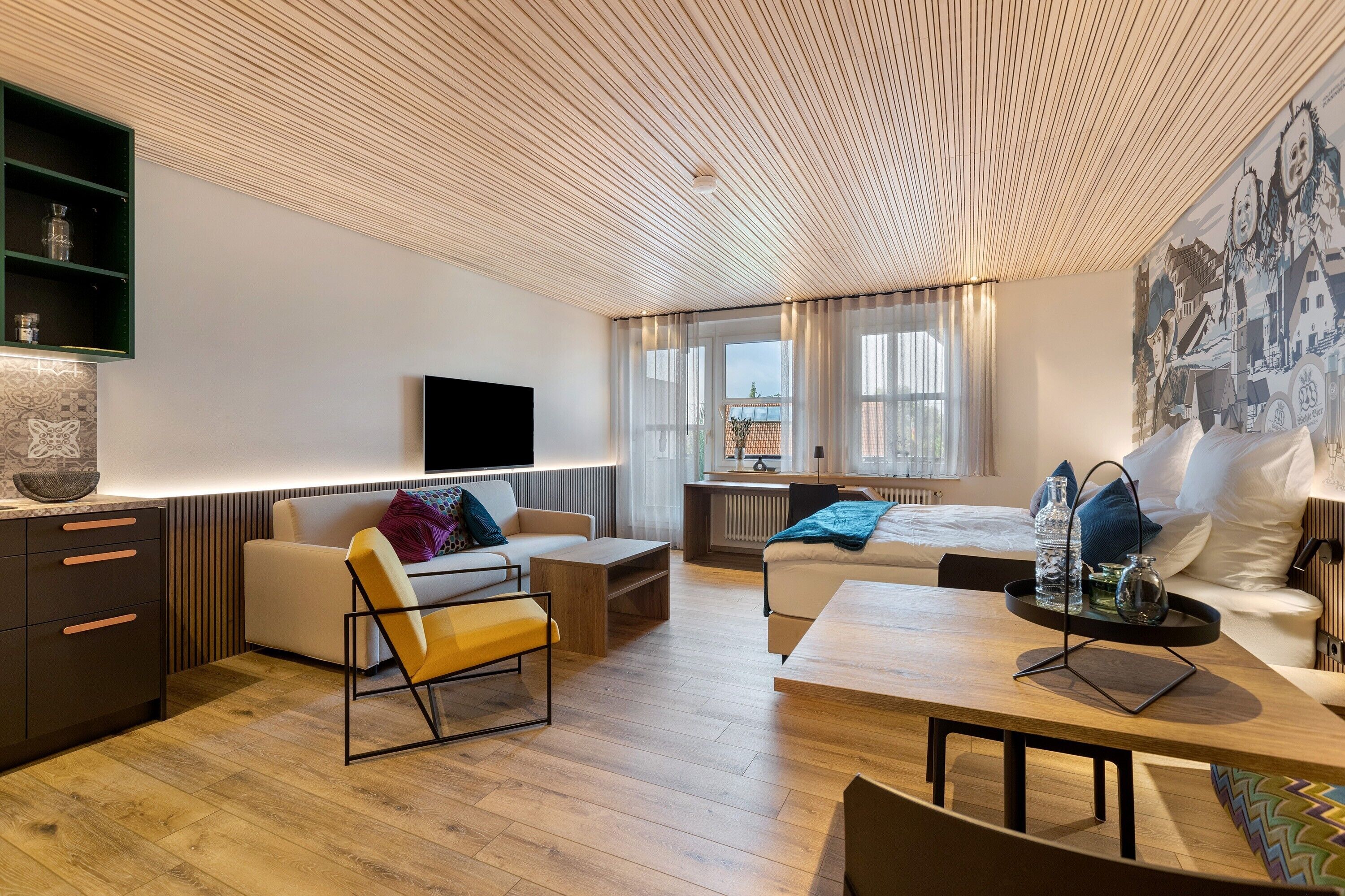 Ein modernes Hotelzimmer mit Holzboden, einem Flachbildfernseher, einem Bett, einer Couch, einem Esstisch und Blick auf die Stadt durch große Fenster.