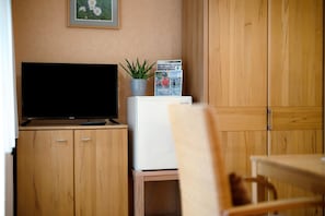 Huoneessa on puukaappeja, televisio, pieni jääkaappi, viherkasvi ja seinällä oleva kehystetty kuva.